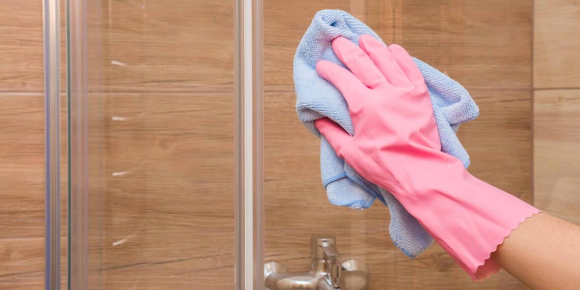 A hand wearing gloves is cleaning a shower glass