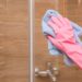 A hand wearing gloves is cleaning a shower glass